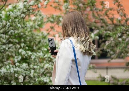 La fille dans la veste sur la rue. Une fille marche dans le parc. Un étranger regarde son téléphone. Banque D'Images