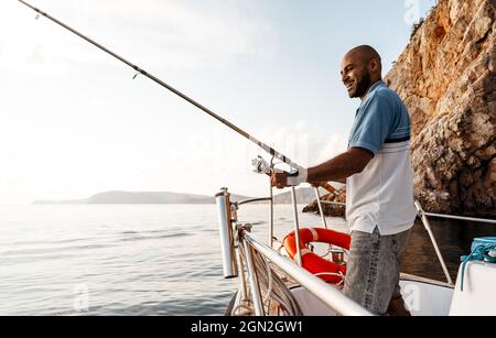 Jeune afro-américain debout avec une canne à pêche sur un voilier pêchant en pleine mer au coucher du soleil Banque D'Images