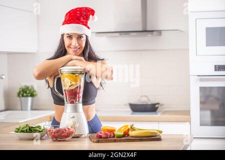 Femme dans le chapeau du Père Noël se penche contre un mélangeur avec un mélange coloré de fruits prêts à être transformé en smoothie. Banque D'Images