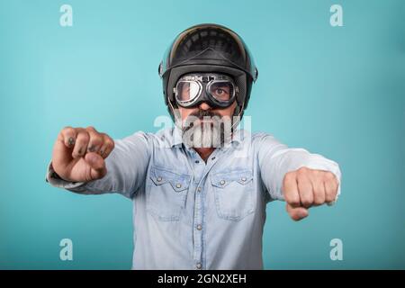 Portrait d'un hipster barbu en chemise denim avec casque et lunettes prétendant monter à bord d'une moto, isolé sur un mur bleu de studio. Banque D'Images
