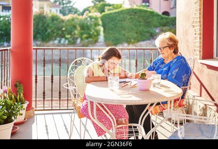 Grand-mère souriante aidant petite-fille gaie à dessiner sur du papier tout en passant du temps ensemble sur la terrasse par beau temps Banque D'Images