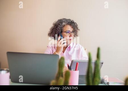 Femme entrepreneure âgée concentrée en lunettes parlant sur un téléphone portable contre un netbook sur une table dans un espace de travail Banque D'Images