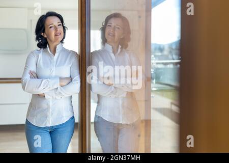 Femme mature et confiante debout regardant la caméra avec les bras croisés s'inclinant sur la porte en verre regardant la caméra le matin à la maison Banque D'Images