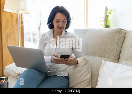 Bonne femme d'âge mûr freelance naviguant sur un smartphone tout en travaillant sur un ordinateur portable assis dans un canapé à la maison Banque D'Images