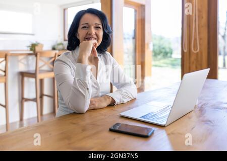 Une femme freelance mûre et heureuse navigue sur Internet sur un netbook en travaillant sur un nouveau projet tout en étant assise à la table à la maison Banque D'Images