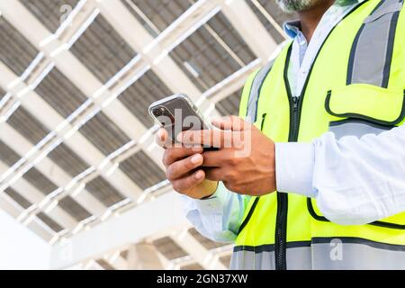 Contremaître court non reconnaissable en gilet de navigation sur smartphone tout en se tenant près de la station d'énergie solaire Banque D'Images