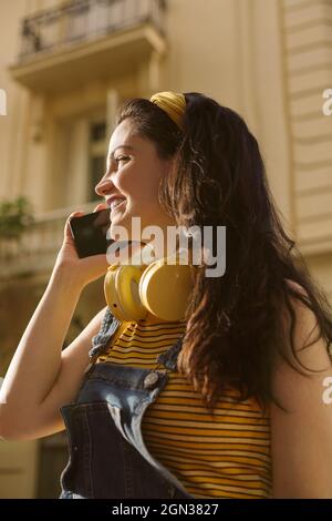 Faible angle de la femelle positive avec casque sans fil sur le cou ayant un appel téléphonique sur le téléphone cellulaire Banque D'Images