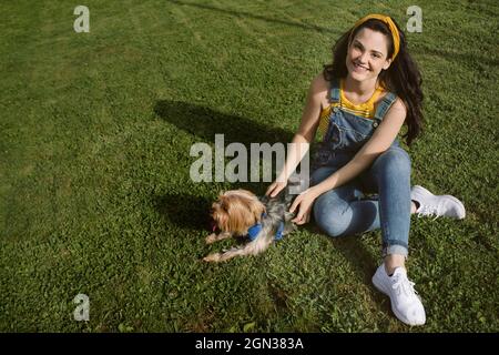 Corps entier à angle élevé de jeune femme heureuse assise sur une pelouse avec des jambes croisées et un Yorkshire Terrier en train de se tasser tout en regardant l'appareil photo Banque D'Images