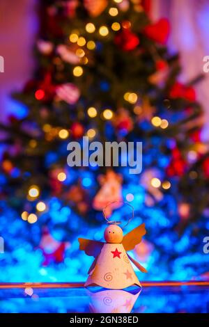 Jouet fait main en forme d'ange placé sur une table réfléchissante contre un arbre de Noël festif avec des guirlandes lumineuses Banque D'Images