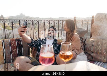 Positif jeune élégant couple assis sur un canapé et de clinking verres de cocktail tout en prenant autoportrait sur le téléphone portable en terrasse à Cappadoce, Turk Banque D'Images