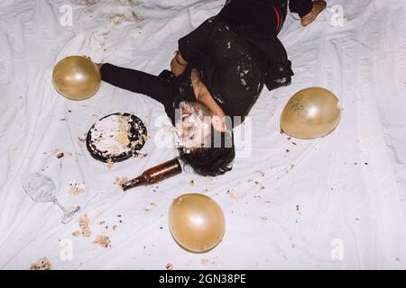 Un homme ivre riant dans un gâteau d'anniversaire écrasé à proximité de bouteilles vides de bière et de ballons avec les yeux fermés Banque D'Images