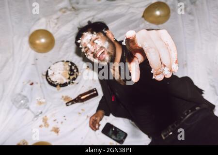 Un homme ivre riant dans un gâteau d'anniversaire écrasé allongé près de bouteilles vides de bière et de ballons et pointant vers l'appareil photo Banque D'Images