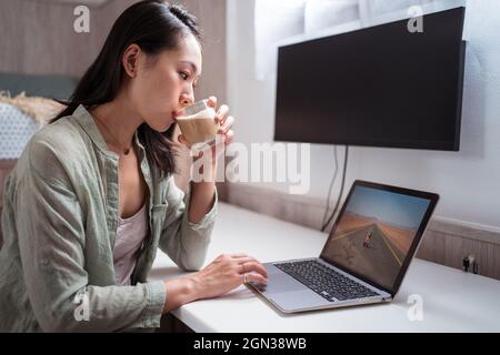 Jeune femme ethnique attentive appréciant le café tout en jouant à un jeu vidéo sur netbook à un bureau dans la maison Banque D'Images
