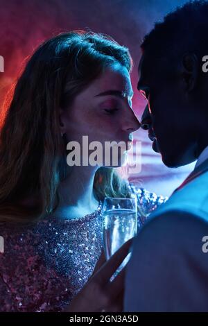 Vue latérale d'un petit couple avec verre de champagne au moment du baiser contre un rayon de lumière brillant regardant l'un l'autre pendant la fête Banque D'Images