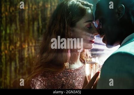 Vue latérale d'un petit couple avec verre de champagne au moment du baiser contre un rayon de lumière brillant regardant l'un l'autre pendant la fête Banque D'Images