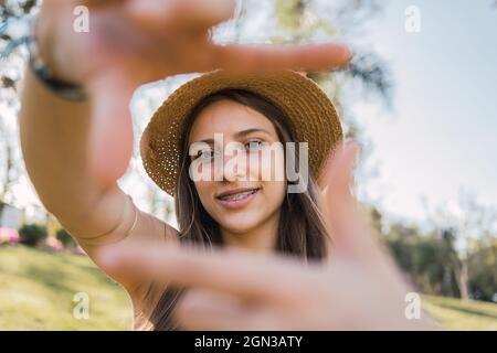 Femme adolescente souriante avec des bretelles démontrant le geste de la photographie tout en regardant l'appareil photo dans la journée sur un arrière-plan flou Banque D'Images