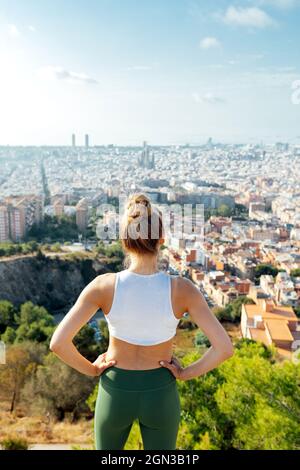 Vue arrière d'une athlète féminine anonyme avec les mains à la taille, en admirant la ville d'été au soleil Banque D'Images