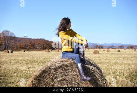 Jeune femme brune en chandail jaune et bottes en caoutchouc assis sur le dessus de haystack dans le champ Banque D'Images
