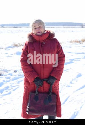 Russe femme âgée de 67 ans, en manteau rouge, regardant la caméra contre un terrain enneigé. Hiver Banque D'Images