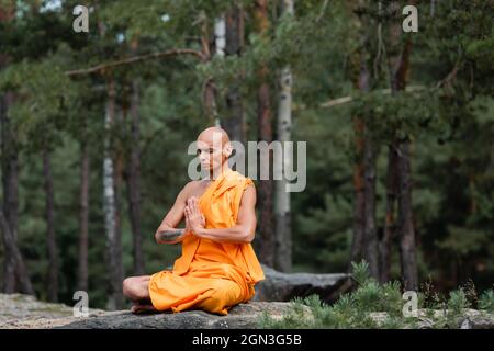 moine bouddhiste dans la kasaya orange assis dans le lotus poser avec les mains de prière tout en méditant dans la forêt Banque D'Images