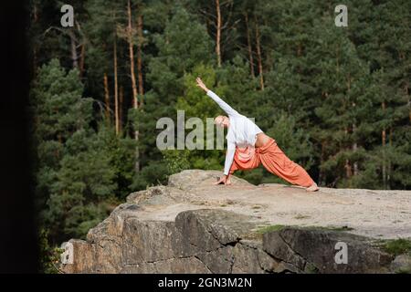 bouddhiste en sweat-shirt et pantalon de harem pratiquant le yoga en fente latérale sur la falaise rocheuse Banque D'Images