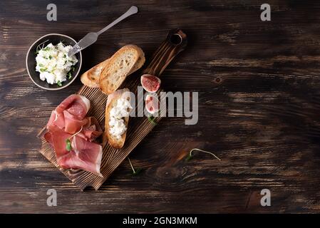 Planche à découper avec prosciutto, tranches de pain et ricotta sur fond de bois foncé, jambon italien avec figues, espace copie. Banque D'Images