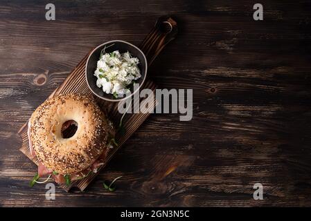 Jambon sec et bagel à la ricotta pour le petit déjeuner sur fond de bois foncé, sandwich avec du fromage prosciutto et des microverts, vue du dessus. Banque D'Images