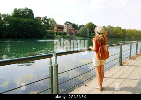 Pleine longueur de fille de voyageur avec sac à dos et chapeau regardant le château du Moyen âge dans le parc. Jeune femme détendue sur la promenade du po à Turin, Italie. Banque D'Images