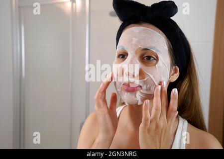 Belle jeune femme applique un masque de tissu cosmétique sur le visage dans sa salle de bains. Concept de soins de santé et de beauté. Banque D'Images