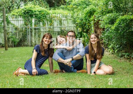 Une famille d'une mère et d'un père et de deux filles assises à l'extérieur dans l'herbe de leur arrière-cour avec la plus jeune fille embrassant papa de derrière Banque D'Images