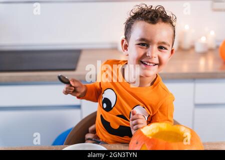 Portrait d'un garçon heureux regardant l'appareil photo habillé comme un t-shirt de citrouille d'halloween vidant et décorant une citrouille d'halloween dans la cuisine à la maison. Banque D'Images