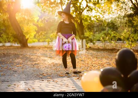 Bonne petite fille fête Halloween en plein air. Enfant en costume de sorcière et chapeau avec ballons orange et noir dans le parc d'automne. Tours pour enfants ou Banque D'Images