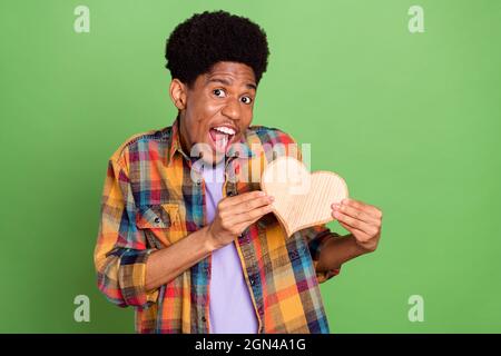 Photo de doux impressionné peau sombre gars habillé damier chemise tenant coeur en bois souriant isolé couleur verte fond Banque D'Images