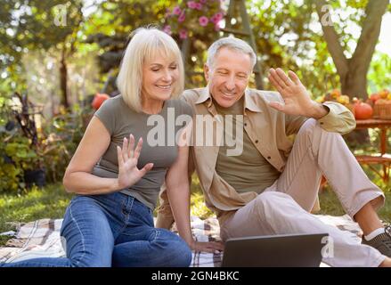 Heureux couple aîné marié utilisant un ordinateur portable et la famille d'appel vidéo, assis sur une couverture de pique-nique dans le jardin. Grands-parents enthousiastes regardant le lapto Banque D'Images