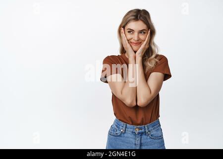 Belle femme blonde serrer son visage et sourire heureux, regardant de côté stupide, debout contre fond blanc Banque D'Images