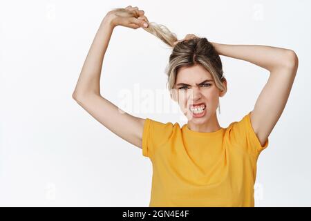 Jeune femme regardant en colère, tirant les cheveux et les dents de serrage, concept de cheveux et produits cosmétiques, debout dans le t-shirt jaune contre blanc Banque D'Images