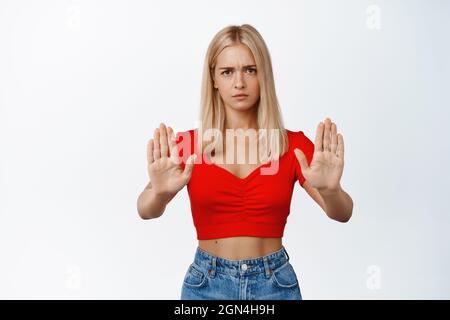 Jeune femme blonde sérieuse montrant un geste d'arrêt, disant non, interdire quelque chose, se tenant sur fond blanc Banque D'Images