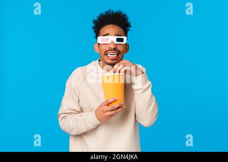 Donc effrayant. Un gars afro-américain terrifié et trémblant regardant un film d'horreur, une scène effrayante dans des lunettes en 3 dimensions, tenant une boîte en papier et Banque D'Images