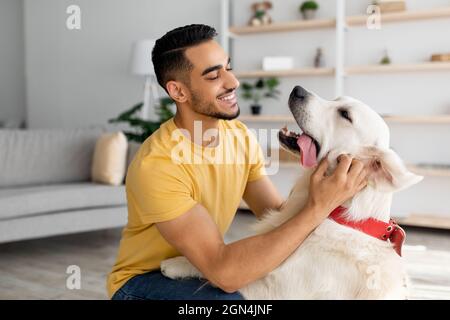 Un jeune homme arabe heureux qui a caressé son adorable chien dans le salon. Concept de l'amitié des animaux humains Banque D'Images