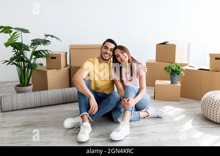 Concept de relocalisation. Heureux propriétaires multiraciaux assis sur le plancher de leur propriété parmi les boîtes en carton, pleine longueur Banque D'Images