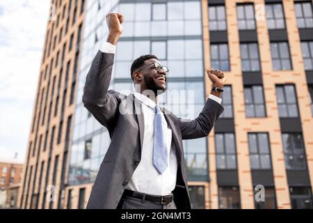 Un homme d'affaires afro-américain enthousiaste qui célèbre son succès et qui se tient debout à l'extérieur près d'un centre de bureaux Banque D'Images