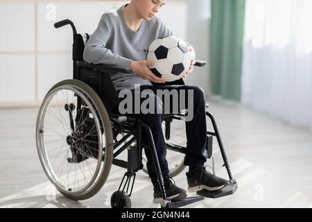 Adolescent handicapé avec ballon de football assis en fauteuil roulant, souffrant de sa blessure, étant incapable de jouer à la maison Banque D'Images