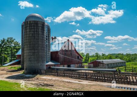 Ancienne ferme rustique avec une grange rouge usée et abîmée et quelques silos entourés d'équipements de ferme d'époque lors d'une journée ensoleillée en été Banque D'Images