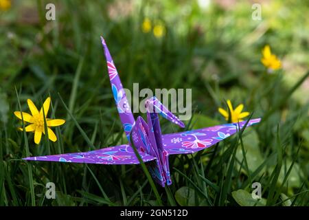 Gros plan d'une grue d'origami en papier, assise sur l'herbe entre les premiers fleurons en pleine floraison, symbolisant le printemps, un nouveau départ et de l'espoir Banque D'Images