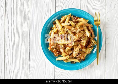 poulet alfredo pâtes penne avec oignon caramélisé arrosé de pignons de pin sur une plaque bleue avec fourchette dorée sur une table en bois blanc, vue horizontale Banque D'Images
