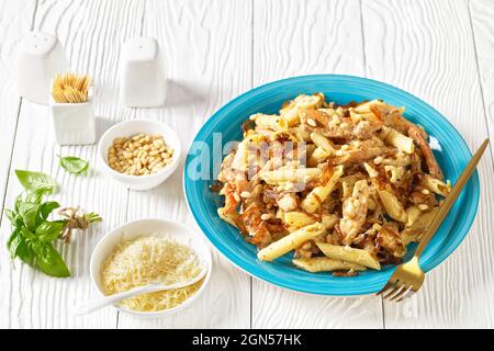 penne de pâtes alfredo de poulet à l'oignon caramélisé arrosé de pignons de pin et de parmesan râpé sur une assiette bleue sur une table en bois blanc, horizont Banque D'Images