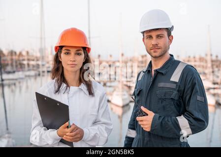 Jeunes professionnels compétents, hommes et femmes, gestionnaires de port en port de protection et casques de sécurité avec presse-papiers et talkie-walkie regardant la caméra en toute confiance en se tenant sur la jetée contre les bateaux amarrés Banque D'Images