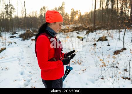 Femme de chemin mobile lors de la course à Woods. Sportswoman avec des bâtons de randonnée tenant le téléphone portable dans les mains le froid jour d'hiver. Banque D'Images