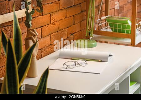 Espace de travail confortable avec ordinateur portable ouvert, crayon et lunettes près du mur de briques Banque D'Images