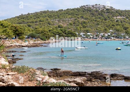 Kosirina, Murter, Croatie - 24 août 2021 : bateaux gonflables amarrés dans une baie calme et des personnes pagayant sur des planches, sur une plage de camping rocheuse Banque D'Images